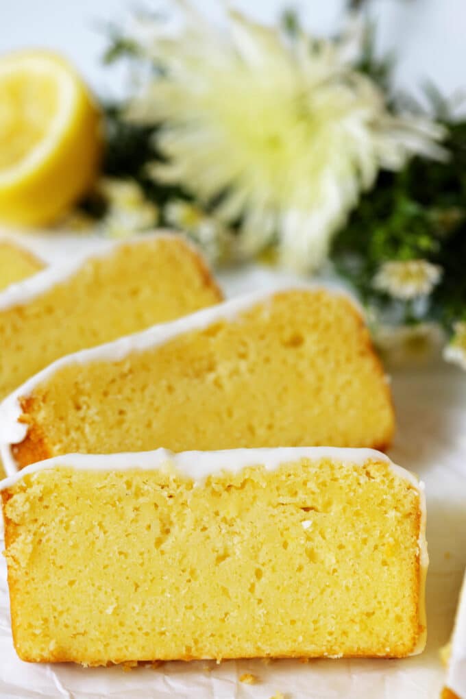 A few vibrant yellow slices of Lemon Pound Cake laying against each other, with a bouquet of flowers laying on the counter behind them.