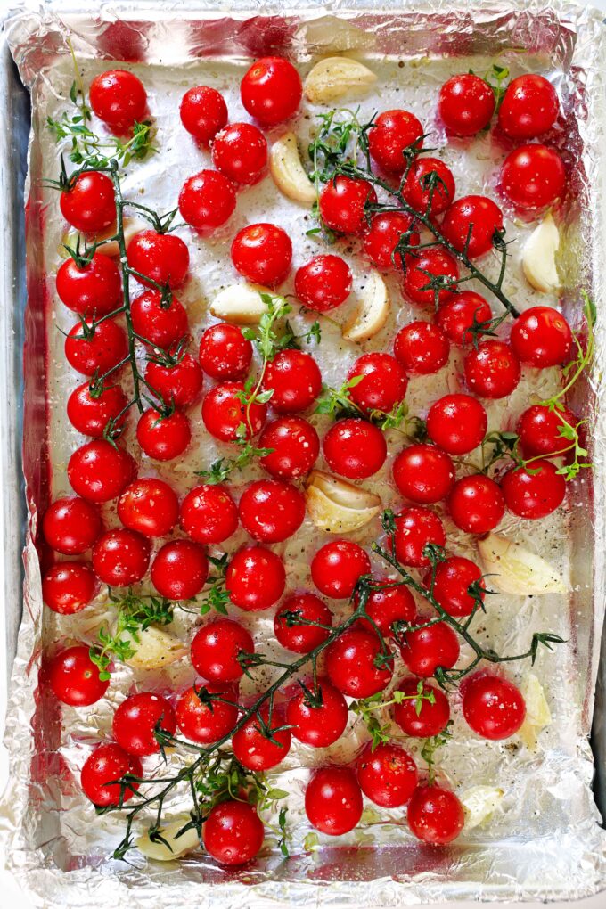 A foil-lined pan of cherry tomatoes, garlic cloves, and sprigs of thyme. Some of the tomatoes are still on the vine.