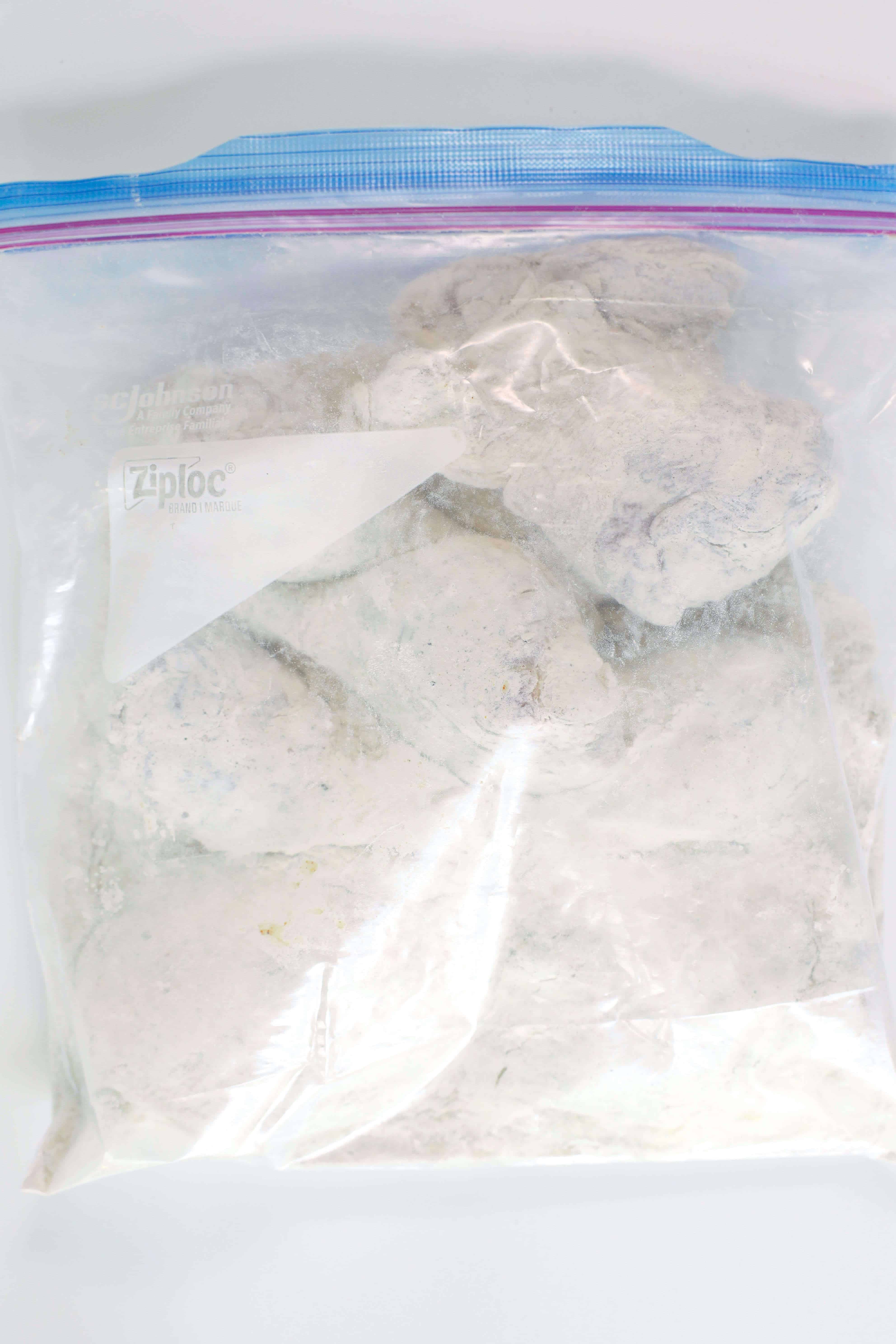 A gallon size zip-top bag full of flour-coated pieces of chicken on a white work surface.