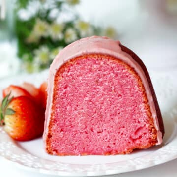 A single piece of Strawberry Pound Cake sitting on a plate with fresh strawberries. The outside is a deep red and the inside is a vibrant pink. There is a light pink glaze drizzled on top.
