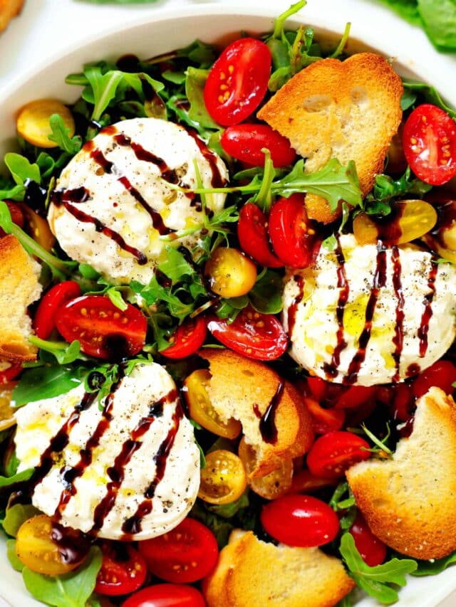 An overhead look at a white bowl full of Burrata Salad. There is arugula, red and yellow tomatoes, burrata, and toasted baguette slices all topped off with olive oil and balsamic glaze, with more bread and basil leaves sitting nearby on the counter.