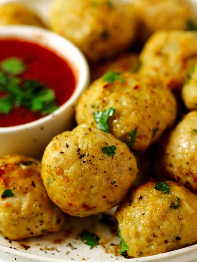 A close up of Chicken Meatballs on a white plate. A bowl of marinara is sitting nearby and there is parsley sprinkled over the meatballs and the sauce.