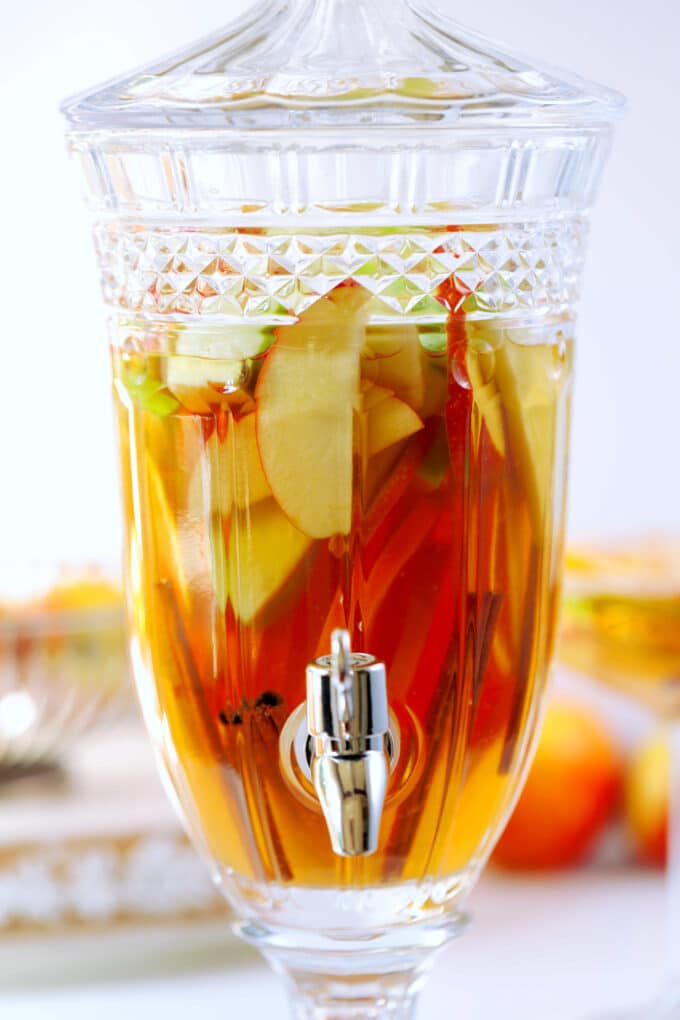 A glass drink dispenser filled with sliced fruit and Apple Cider Sangria.