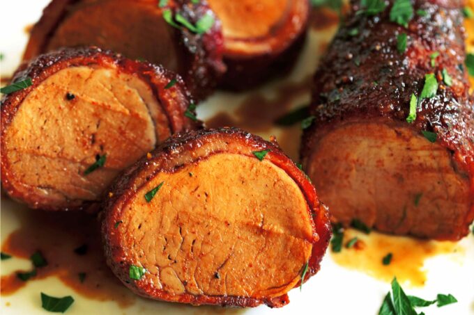 Several Bacon Wrapped Pork Tenderloin medallions leaning against each other on a with plate with a chopped parsley sprinkled on top.