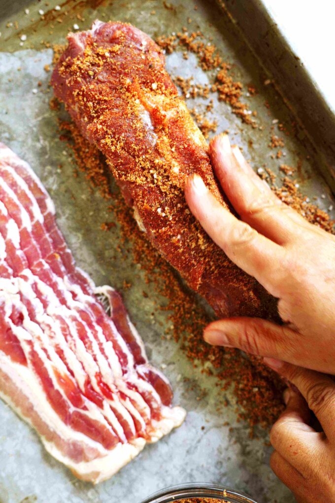 A hand rubbing the brown sugar mixture into a pork tenderloin. A stack of bacon is sitting next to it.