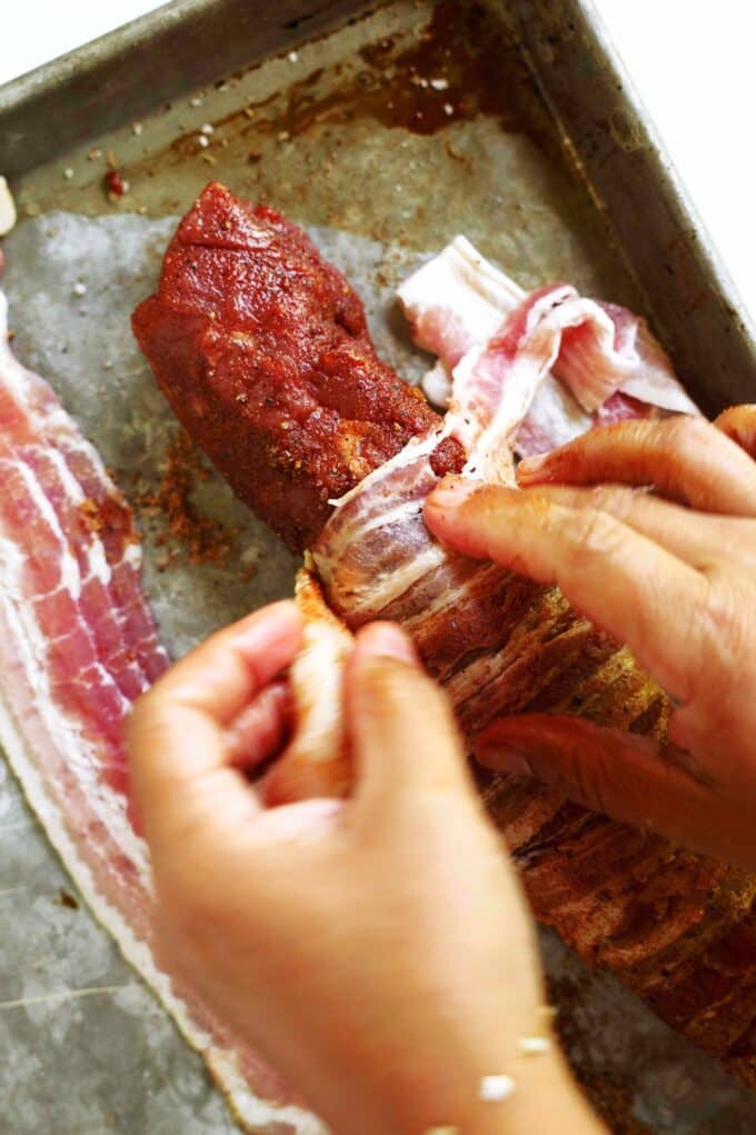 Hands carefully wrapping a pork tenderloin in slices of bacon.
