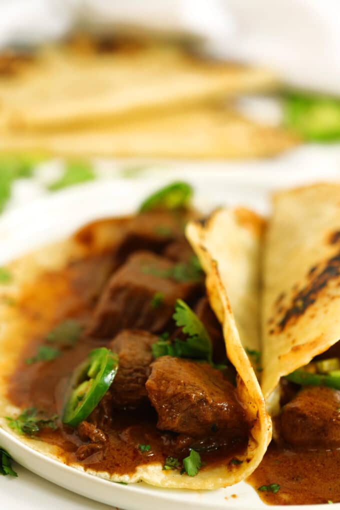 A close up of two Carne Guisada tacos on a white plate with chopped cilantro sprinkled on top.