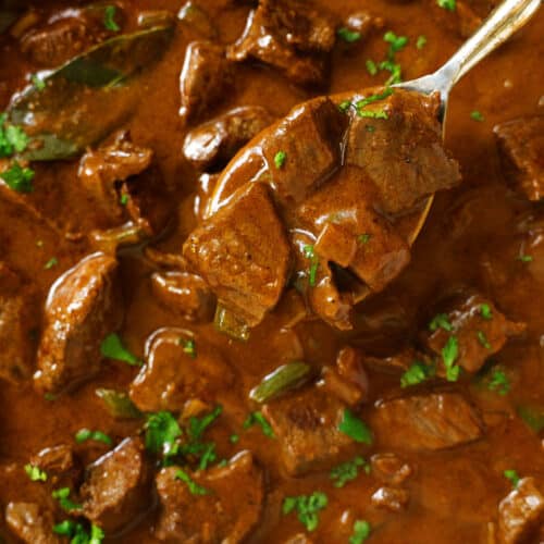 A spoon being used to dip a scoop out of a pan of Carne Guisada. The mixture is dark brown with flecks of bring green cilantro.