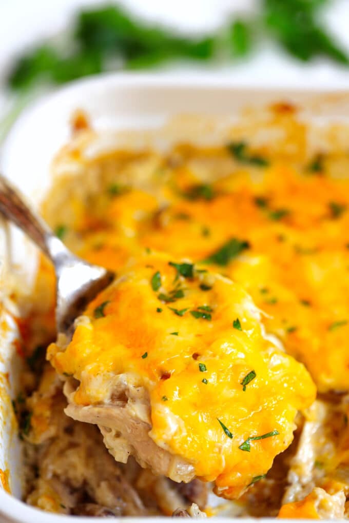 A spoon lifting a scoop of Chicken and Rice Casserole out of the pan. Melty, yellow cheese, shredded chicken, cooked rice and mushrooms are all visible.