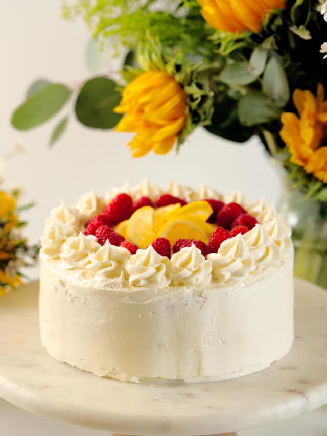 A whole Lemon Raspberry Cake sits on a cake stand. It has frosting piped around the top with lemon wheels and fresh raspberries in the middle. There is a vase full of sunflowers and greenery behind it.