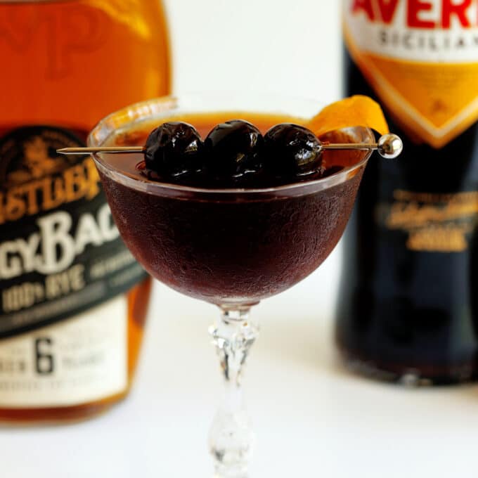 A Black Manhattan in a coupe glass with three Luxardo maraschino cherries on a skewer across the top, and an orange twist on the side. There are bottles of whisky in the background.