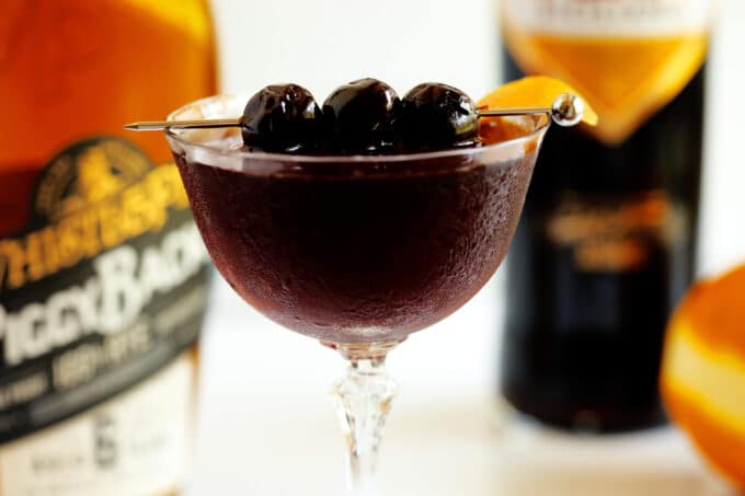 A close up of a Black Manhattan in a coupe glass with three Luxardo maraschino cherries on a skewer across the top, and an orange twist on the side.