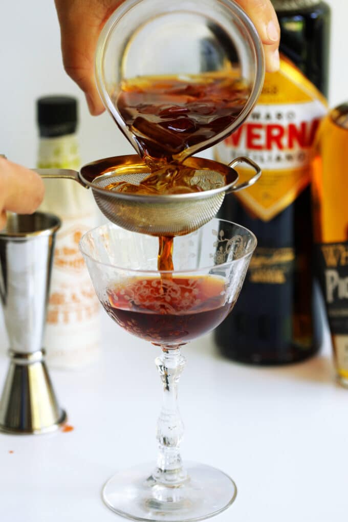 Black Manhattan ingredients being poured through a strainer into a coupe glass with bottles of ingredients sitting in the background.