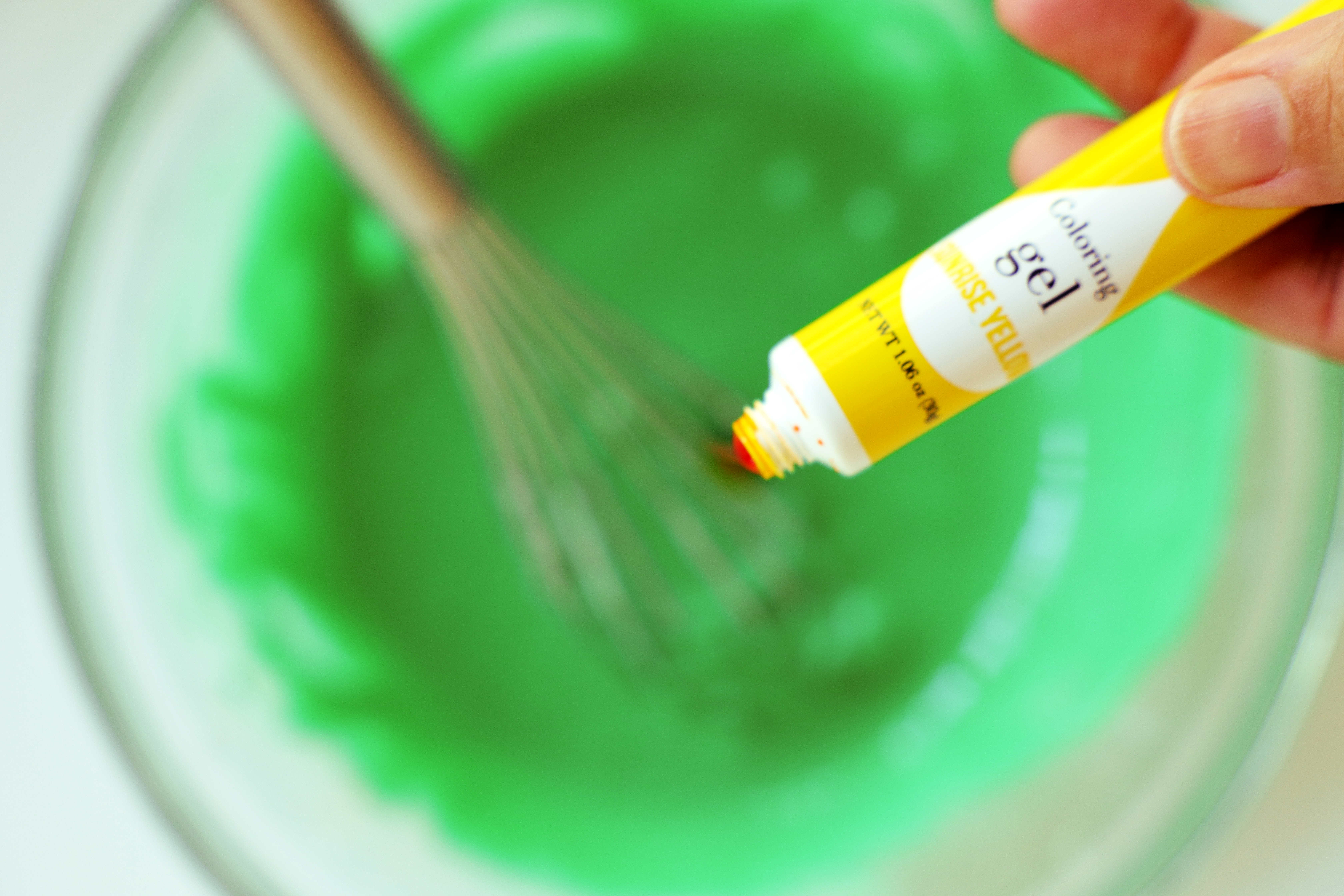 Large, clear mixing bowl containing green icing and a silver whisk. Yellow food coloring gel is being held above the bowl.