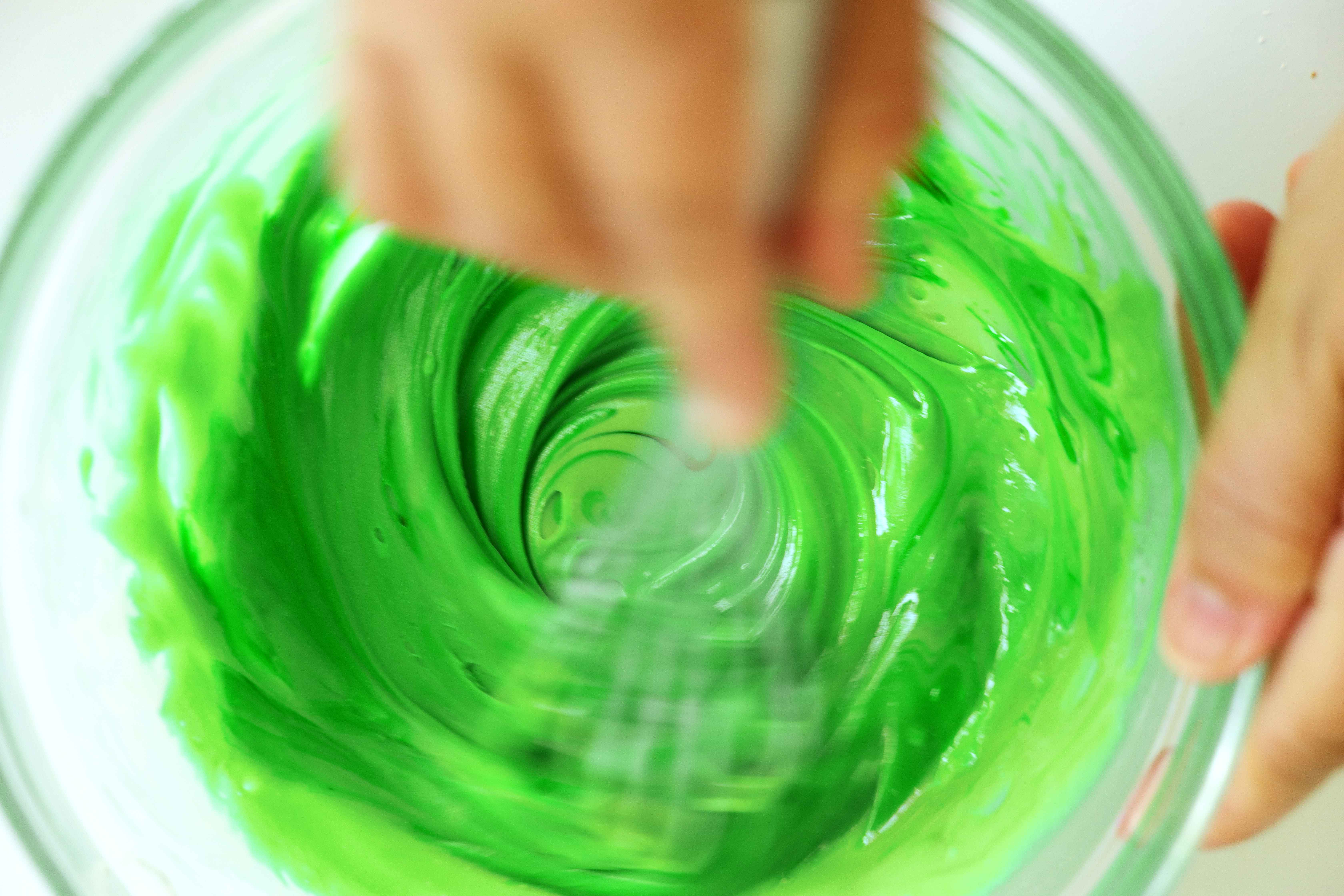 Green icing being whisked in a large, clear mixing bowl.