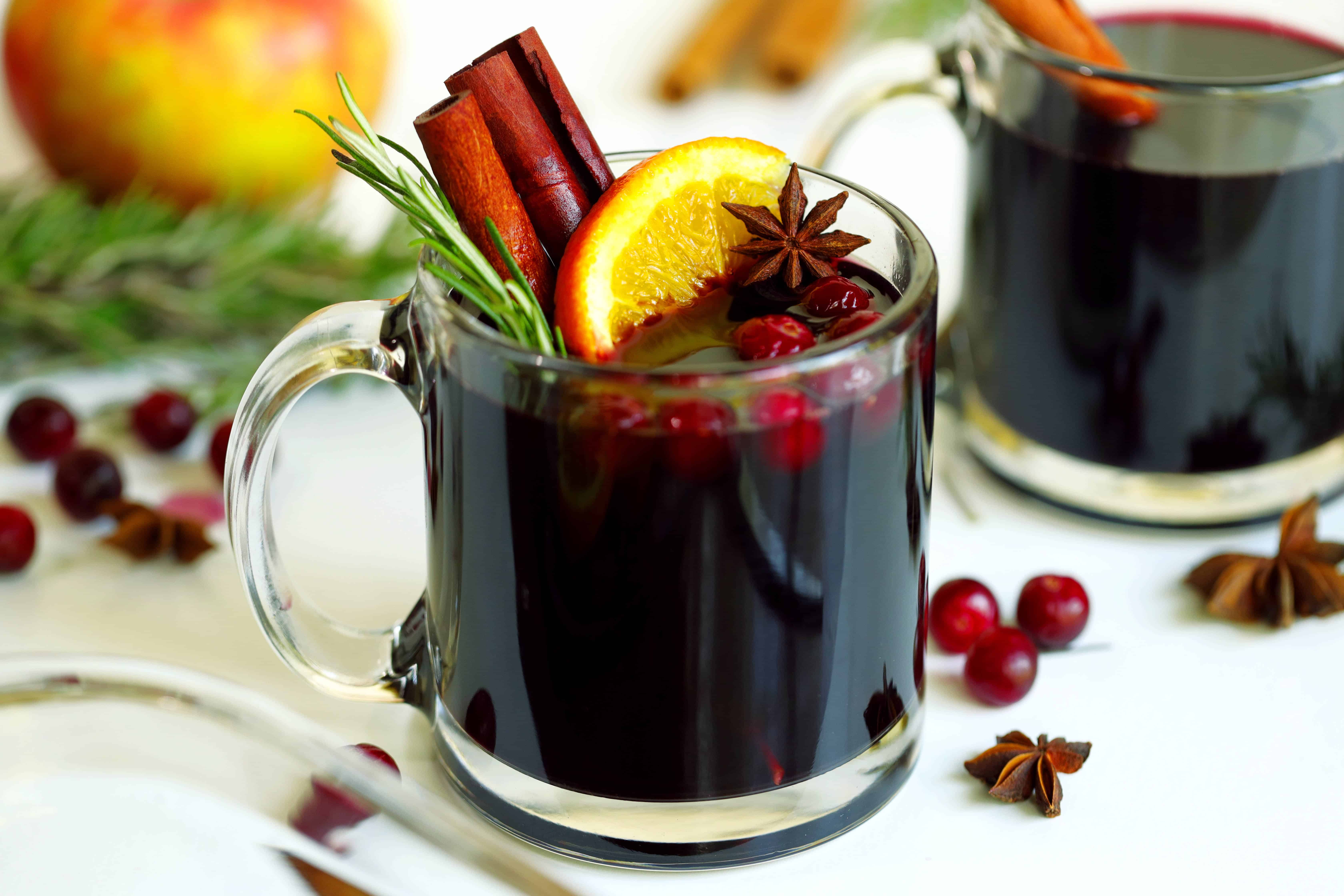 Clear mug on a white table cloth, holding the red Mulled Cider cocktail. Garnished with red cranberries, rosemary sprig, orange slice, cinnamon sticks, and a star anise.