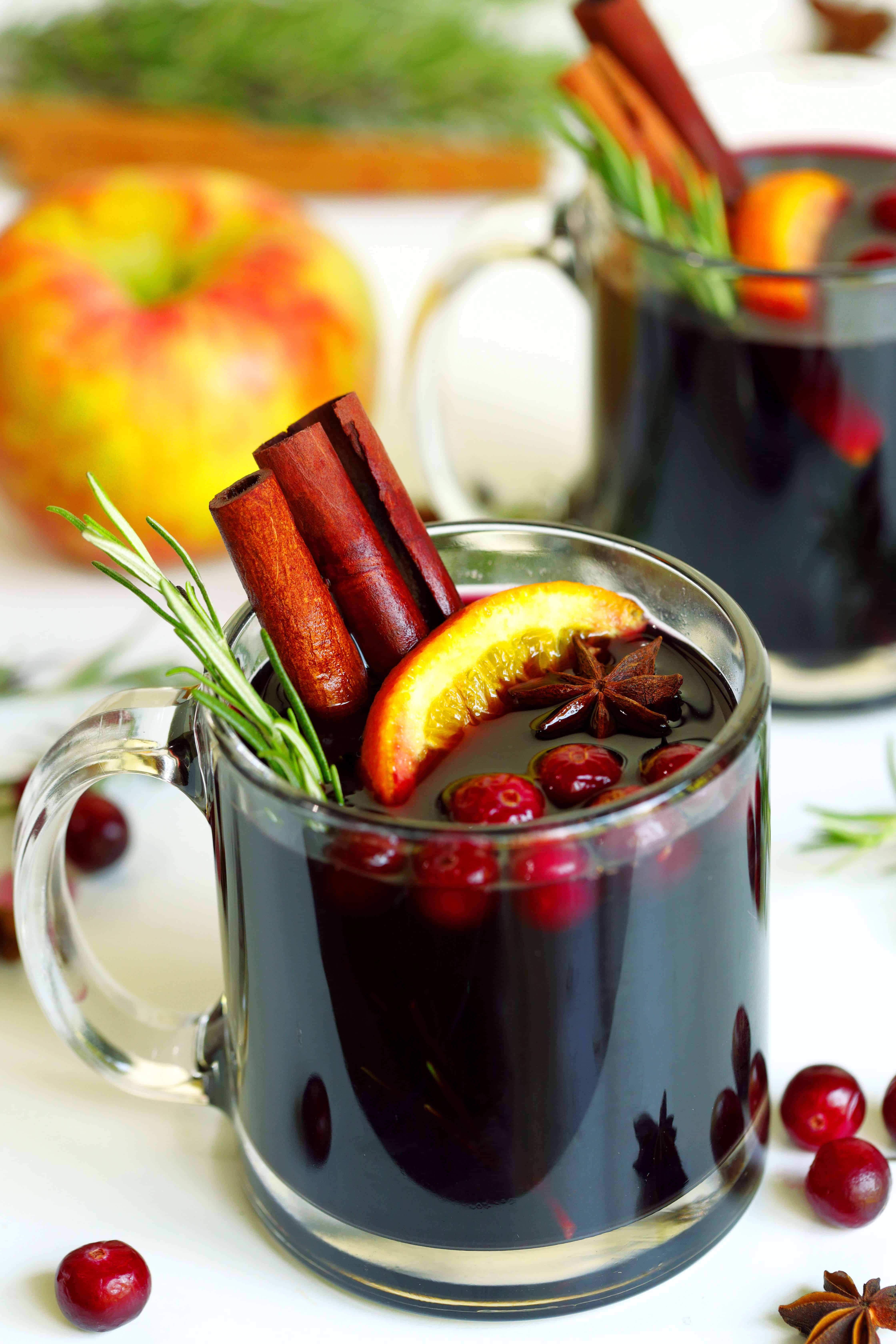 Mulled wine in a clear mug, garnished with rosemary sprig, cinnamon sticks, apple slice, star anise, and cranberries. Mug is sitting atop a white table cloth surrounded by cranberries on the table.