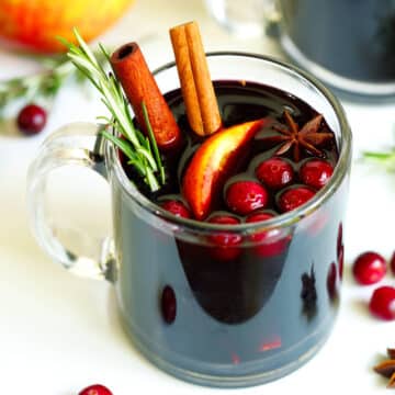 Clear mug with red mulled cider cocktail on a white table cloth with raw cranberries and rosemary sprigs sprinkled around. Garnished with two cinnamon sticks, one apple slice, seven cranberries, a sprig of rosemary, and one star anise.