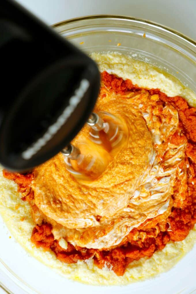 A hand mixer being used to mix pumpkin purée and sour cream into other wet ingredients.