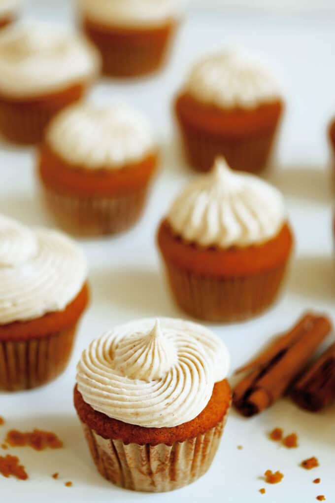 Several Pumpkin Cupcakes with piped, cream cheese frosting sitting on a white surface with some cinnamon sticks laying nearby.