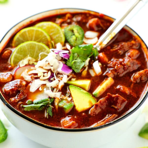 White bowl with black rim containing Pozole Rojo - red broth, chunks of pork, grated cheese, chopped red onion, three lime slices, sliced jalepeno rounds, avocado chunks, and chopped cilantro.