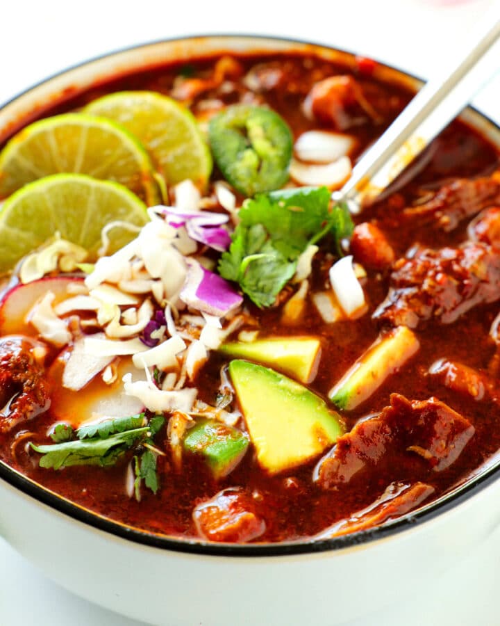White bowl with black rim containing Pozole Rojo - red broth, chunks of pork, grated cheese, chopped red onion, three lime slices, sliced jalepeno rounds, avocado chunks, and chopped cilantro.