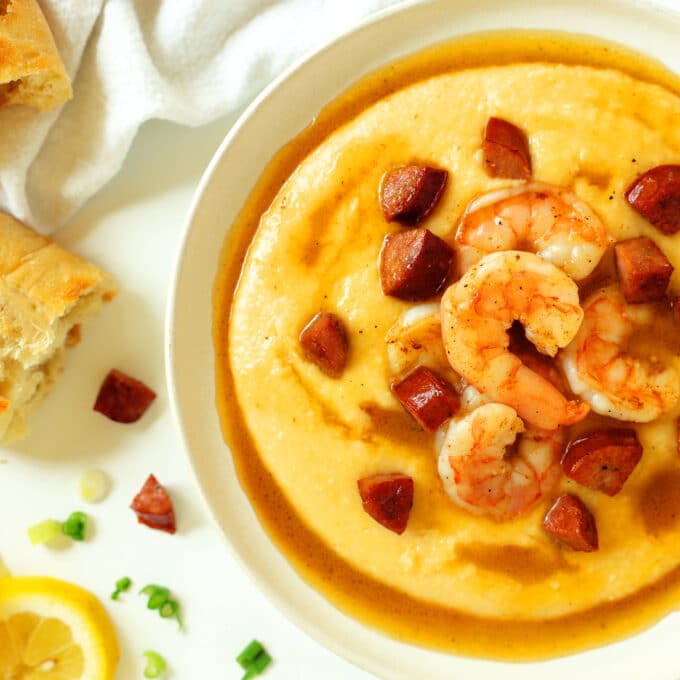 An overhead look at a bowl of grits with sausage, shrimp, and a thin sauce on top. There are pieces of bread sitting nearby.