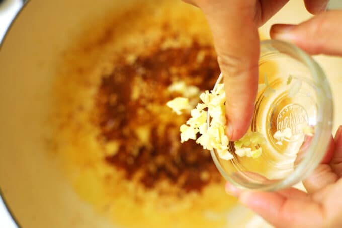 Hands swiping minced garlic from a small glass bowl, into a pan of oil.