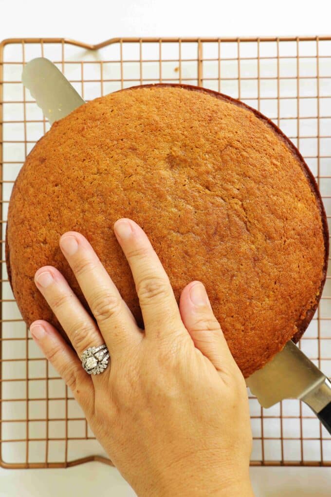 A large, serrated knife being used to cut the dome off the top of a Sweet Potato Cake that is sitting on a cooling rack.