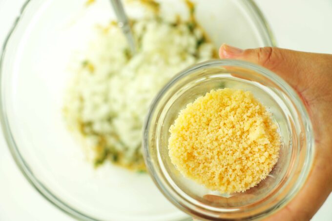 A hand holding a small bowl of grated Parmesan cheese over a bowl of breadcrumbs and herbs.