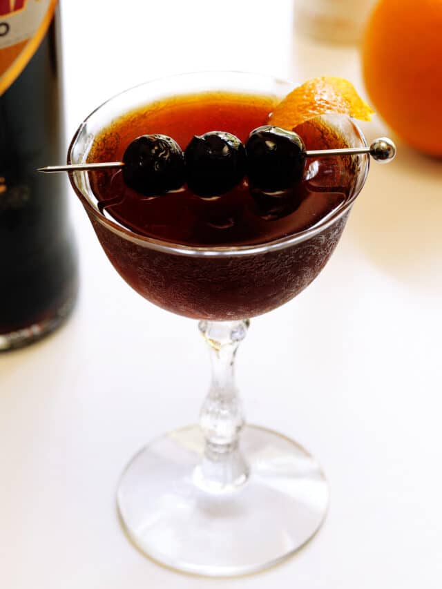 A Black Manhattan in a coupe glass with three Luxardo maraschino cherries on a skewer across the top, and an orange twist on the side.