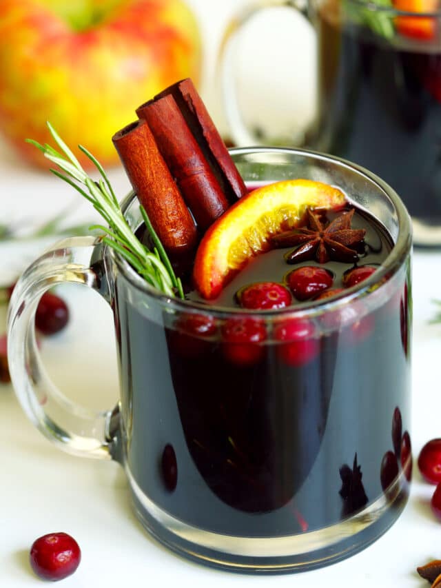 Mulled wine in a clear mug, garnished with rosemary sprig, cinnamon sticks, apple slice, star anise, and cranberries. Mug is sitting atop a white table cloth surrounded by cranberries on the table.
