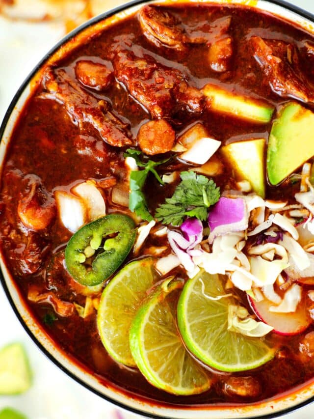 White bowl with black rim containing Pozole Rojo - red broth, chunks of pork, grated cheese, chopped red onion, three lime slices, sliced jalepeno rounds, avocado chunks, and chopped cilantro.
