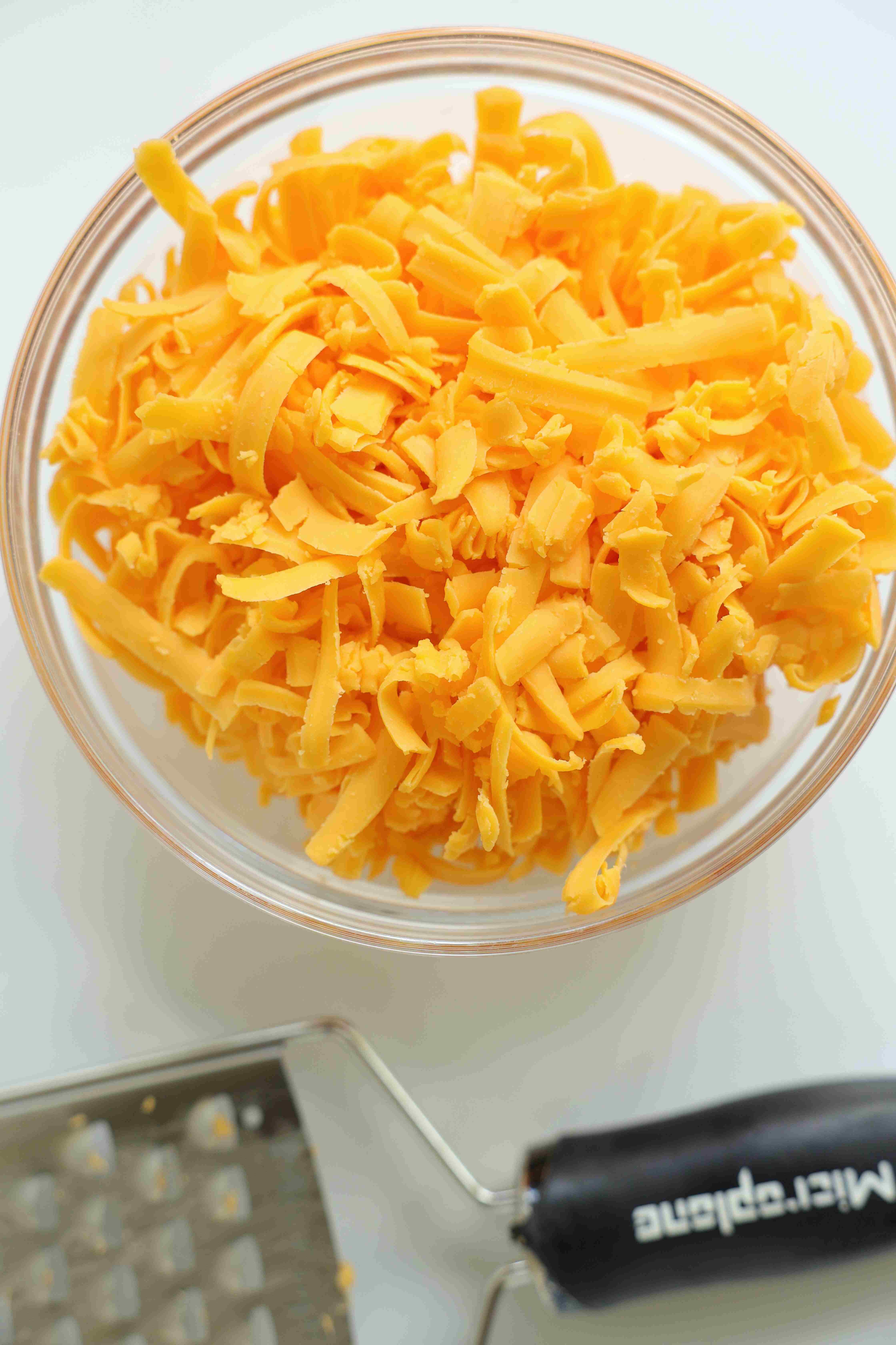 Freshly grated cheddar cheese in a clear mixing bowl next to a microplane tool.