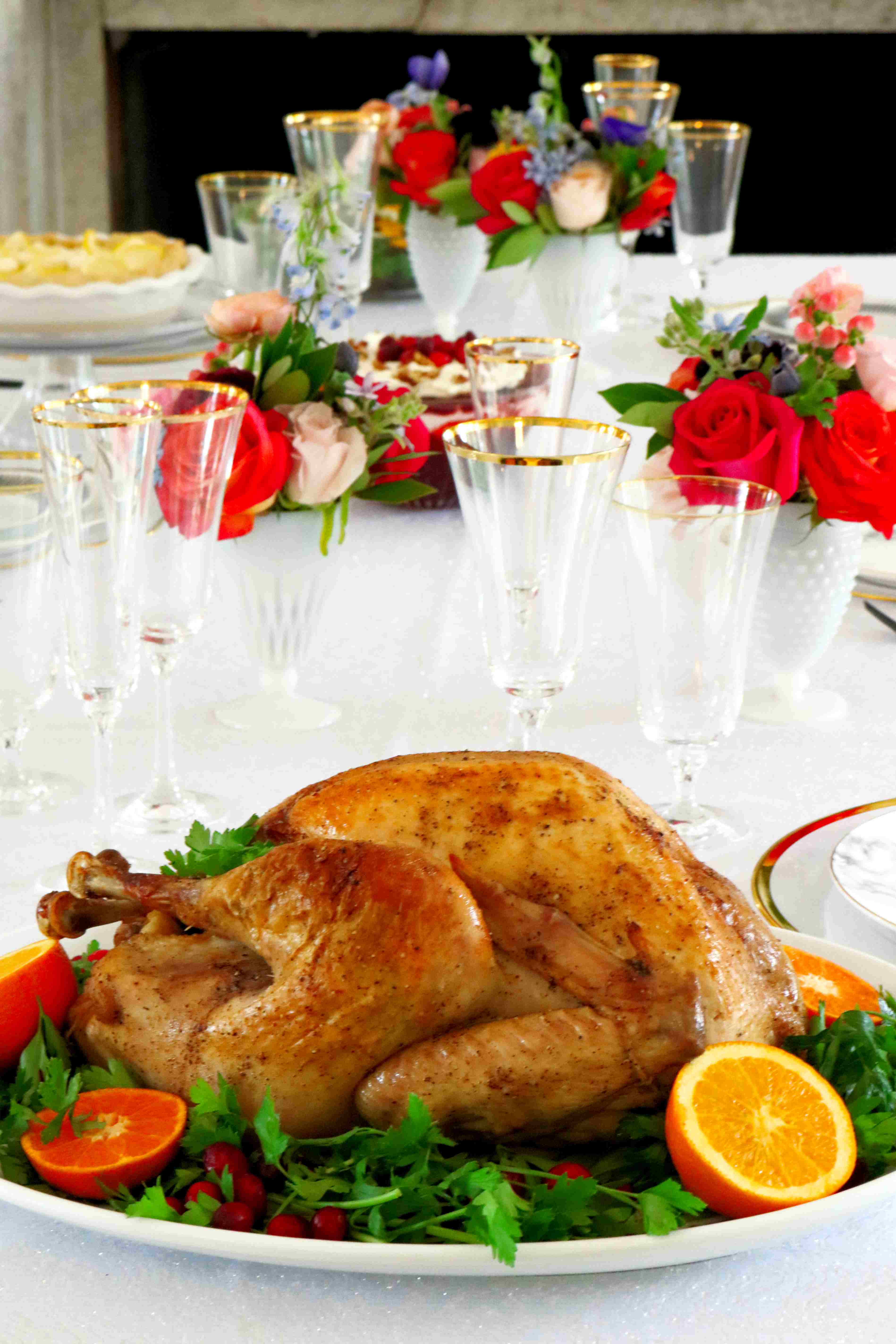 Whole roasted turkey on a platter with parsley and oranges around it. It is on a white tablecloth with flowers in the background.