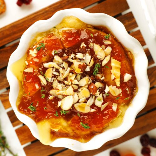Baked Brie with Jam, chopped almonds, fresh thyme, and drizzled honey in a white serving dish, sitting on a wooden trivet