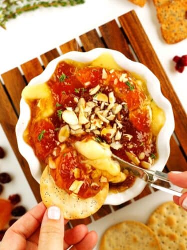 Overhead shot of cheesy baked Brie in a small serving dish topped with preserves, chopped almonds, fresh herbs, and being spread on a thin cracker.