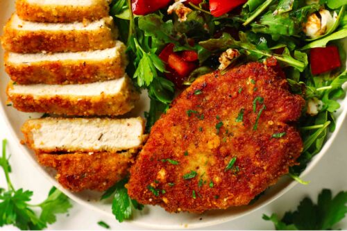 Overhead shot of two Parmesan Crusted Pork Chops, one sliced and whole, with a bed of mixed green, chopped tomatoes, and balsamic vinegar.