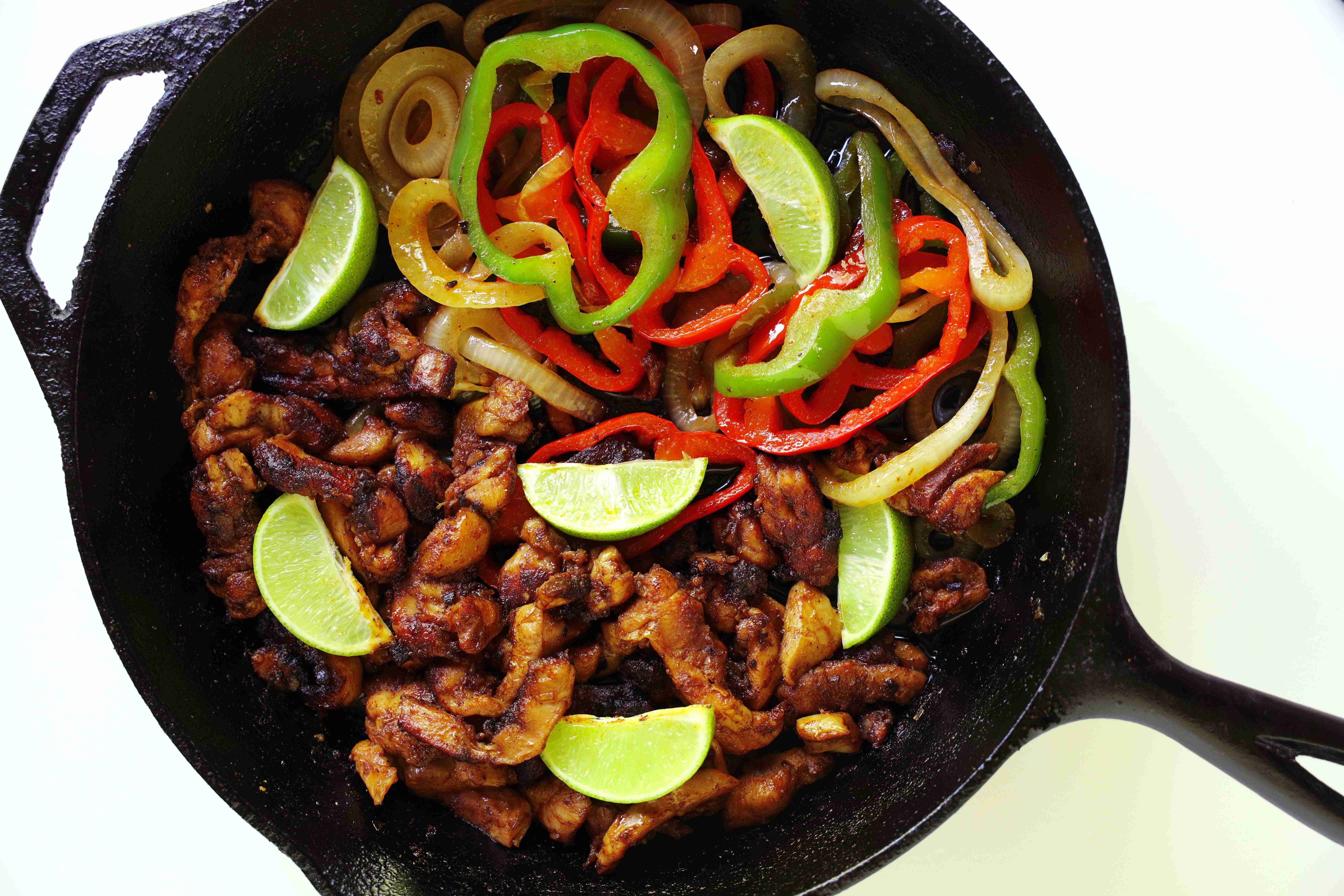 Cooked chicken thighs and sauteed peppers and onions for Chicken Fajita Tacos in a cast iron skillet with slices of lime on top.
