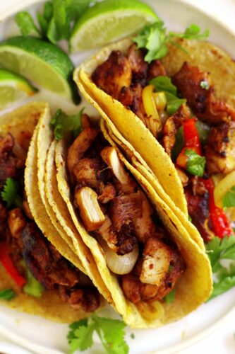 3 chicken fajita tacos on a white plate with lime wedges and cilantro off to the side.