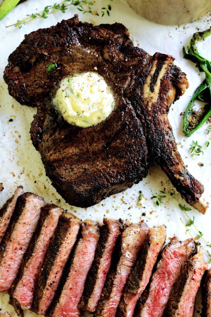 An overhead shot of a cowboy steak with butter on top alongside slices of cowboy steak.