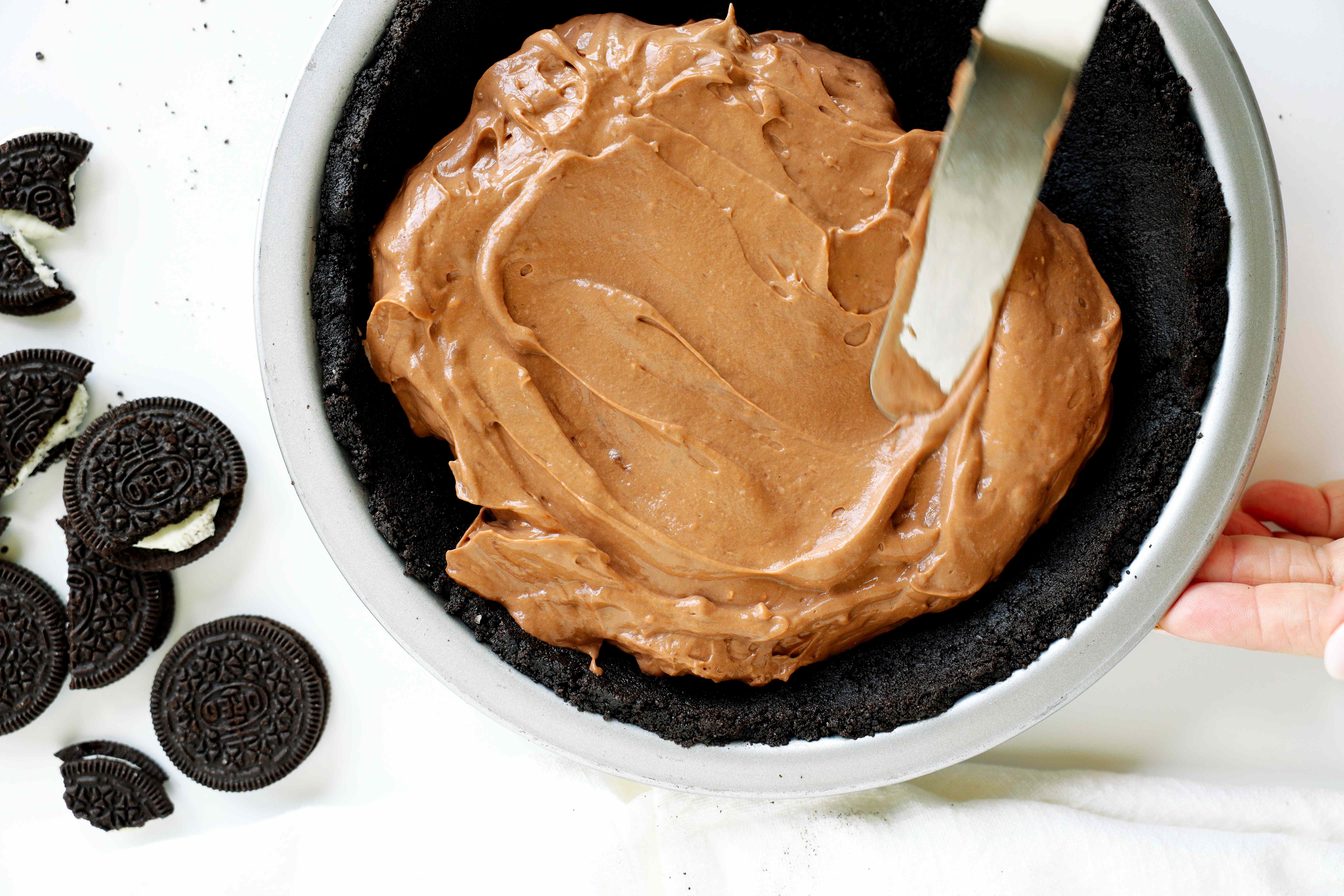 Oreo pie crust with chocolate pudding filling being spread inside. Whole and broken pieces of Oreo sandwich cookies on the side of the pie dish.