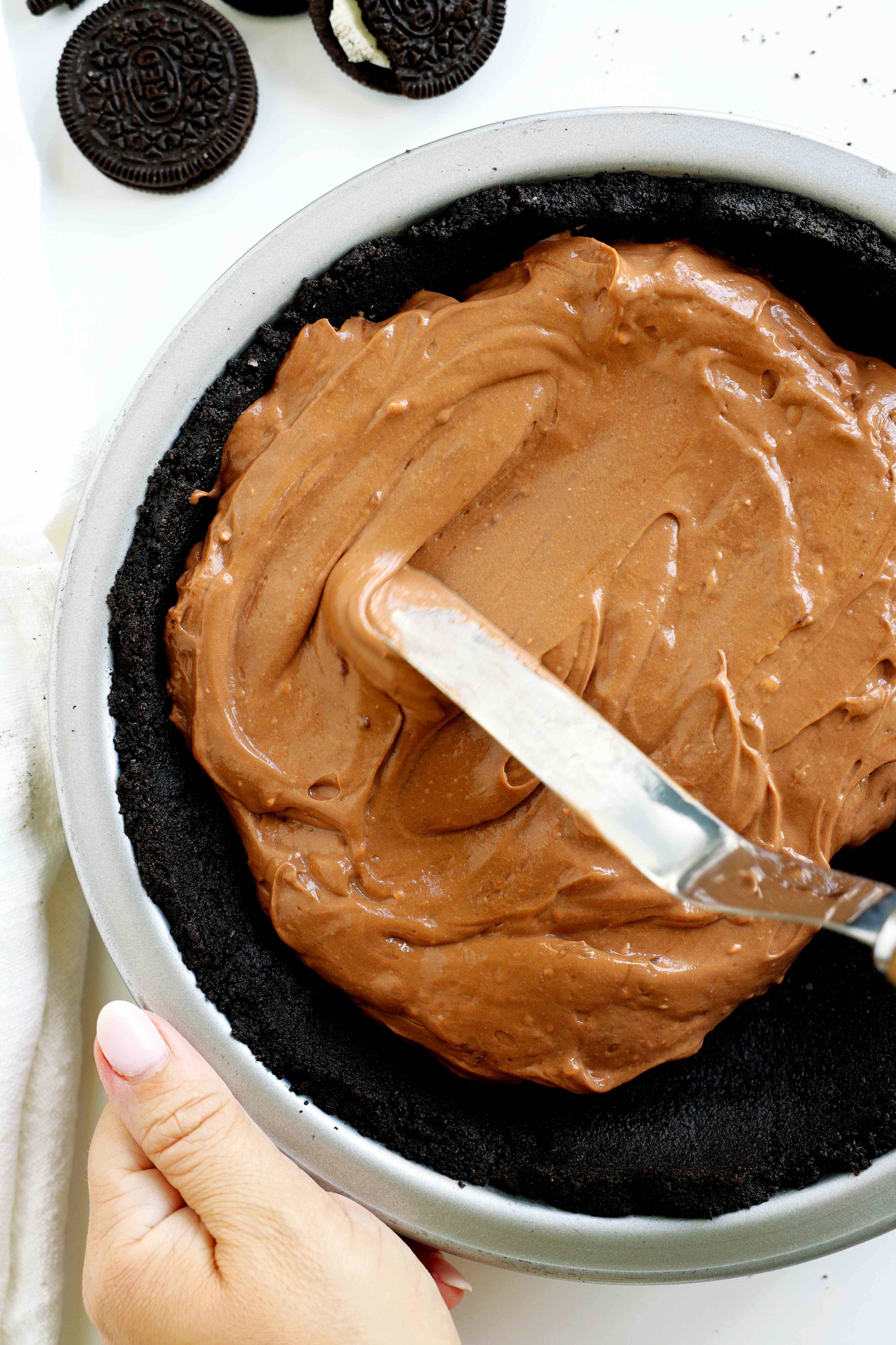 Oreo pie crust with chocolate pudding filling being spread inside. Whole and broken pieces of Oreo sandwich cookies on the side of the pie dish.