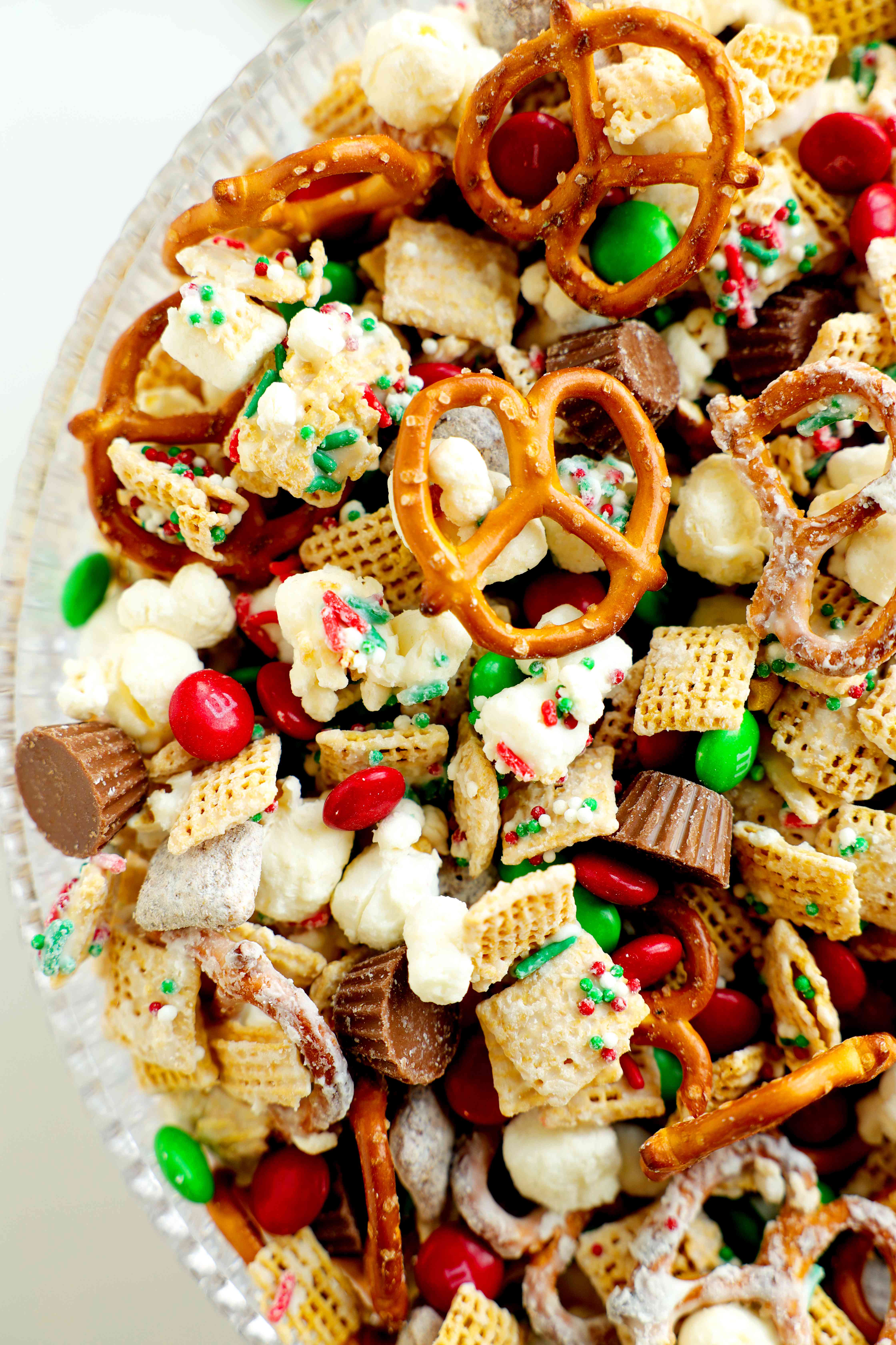 An overhead shot of Christmas snack mix, and a clear glass bowl with pretzels, cereal, marshmallows, Reese's, and M&M's.