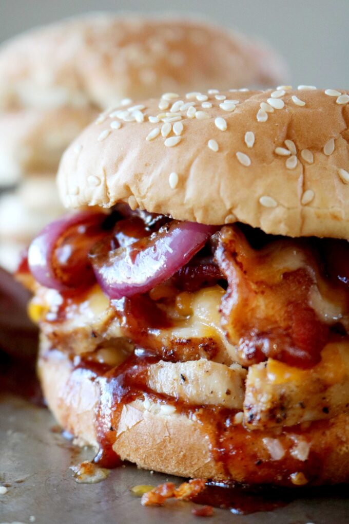 A close-up of a barbecue chicken sandwich on a sesame seed bun, layered with grilled chicken, crispy bacon, melted cheese, caramelized onions, and a drizzle of barbecue sauce.