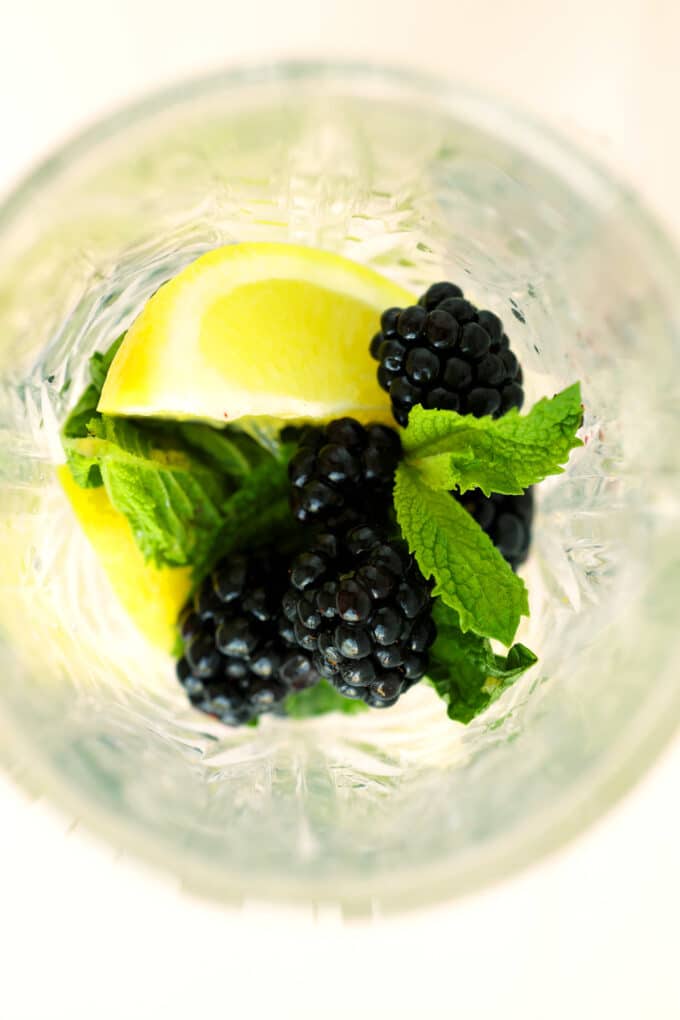 Overhead view of fresh blackberries, bright yellow lemon wedges, and vibrant green mint leaves in a clear cut-glass cocktail shaker.