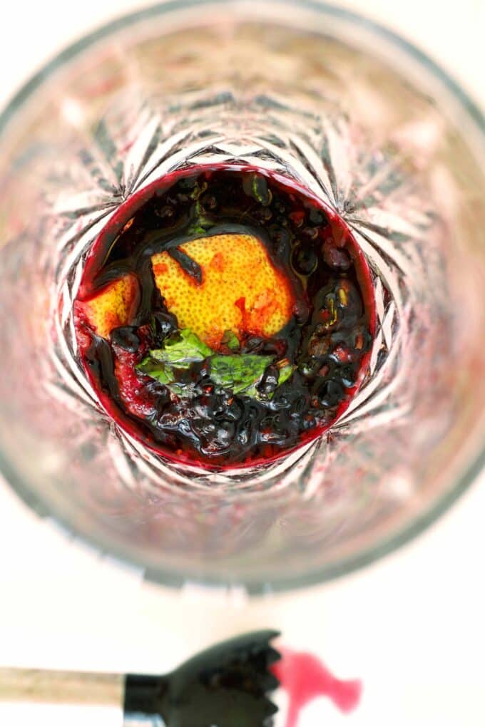Overhead view of muddled blackberries, lemon, and mint in a clear cut-glass cocktail shaker, releasing deep purple juice and citrus oils.