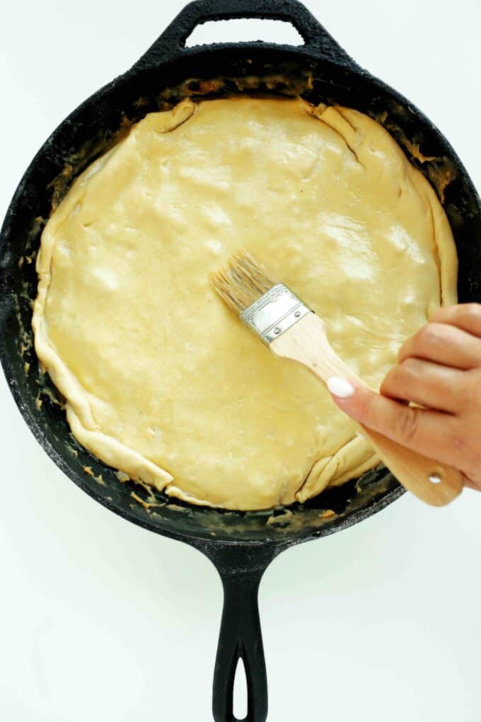 Overhead shot of uncooked Chicken Pot Pie with Puff Pastry in a cast iron skillet, adding butter with a pastry brush