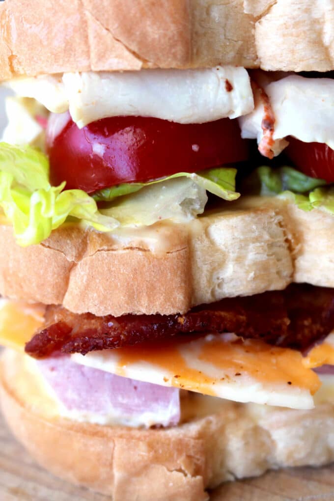 A close-up of a stacked club sandwich on white bread with layers of crisp bacon, cheddar and white cheese, deli turkey, lettuce, tomato slices, and mayonnaise, showcasing the hearty texture and fresh ingredients.