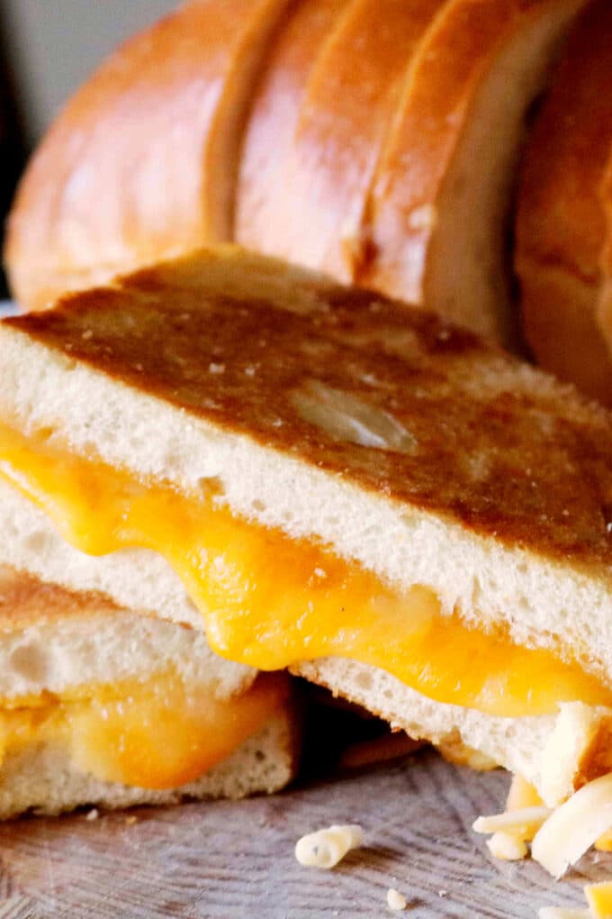 Close-up of a golden, crispy grilled cheese sandwich with melted cheddar oozing between thick slices of toasted bread, with a loaf of bread in the background.